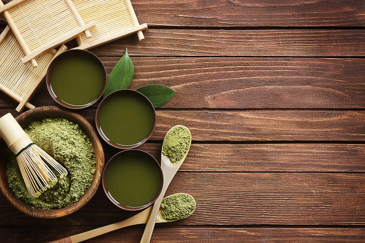 Matchapulver, en bambuvisp och koppar med vispad matcha på ett träbord